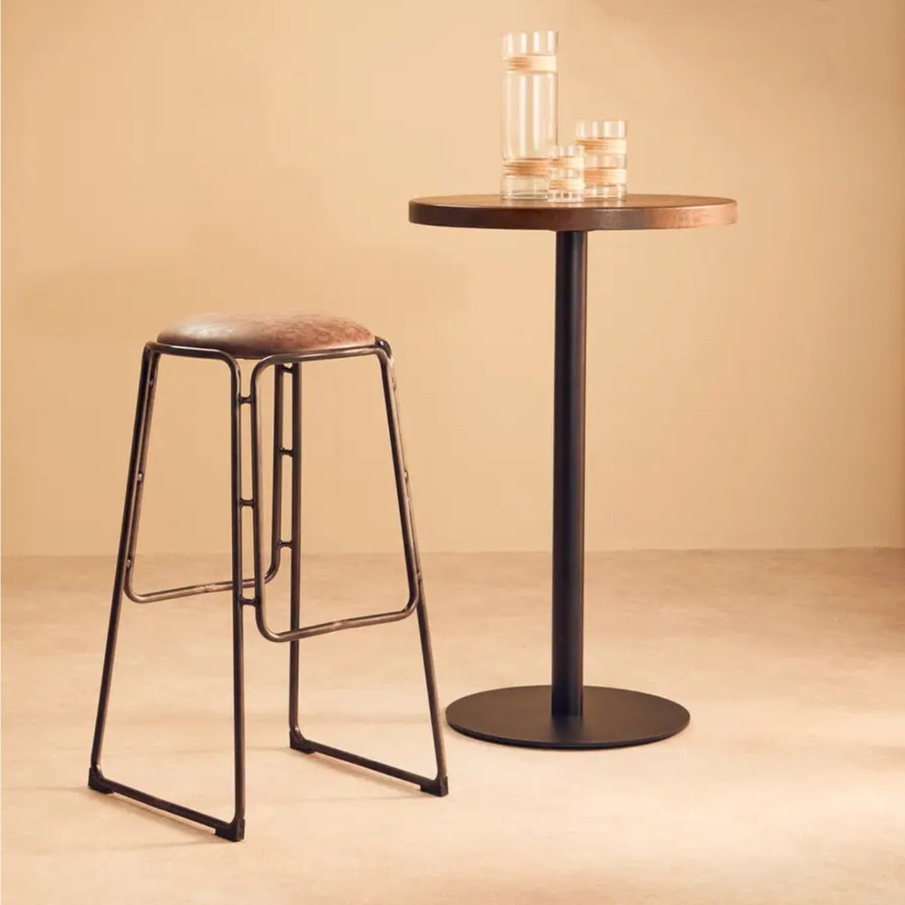 Lifestyle shot of the Dali bar stool next to a tall bar table in a room full of metallic, industrial design elements. the solid soldering of the chair's legs and footrests is on full display.