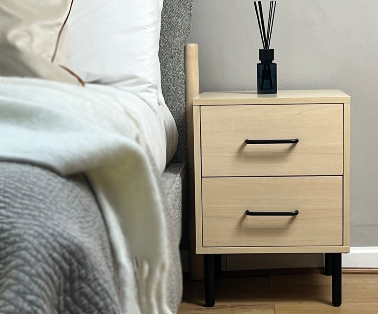 Lifestyles shot of the William 2 drawer bedside next to a modern upholstered bed with grey fabric showing the oak finish of the bedside complementing the modern bed.
