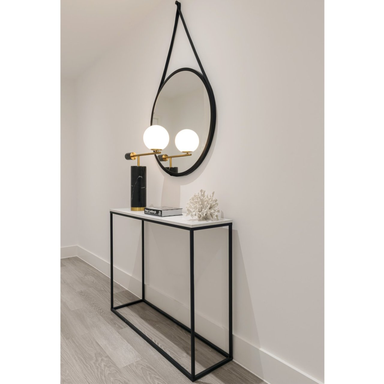 Lifestyle shot of the round mirror with black frame attached in a hallway over a console table with a lamp in front of it dispersing the light throughout the space