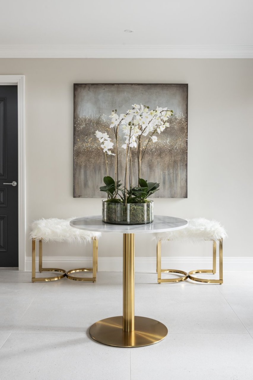 A round dining table with a marble top and sleek brass metal base sits in front of two fluffy stools and an abstract wall art. A plant sitting on top of the table creates a perfect amalgam with the painting behind it.