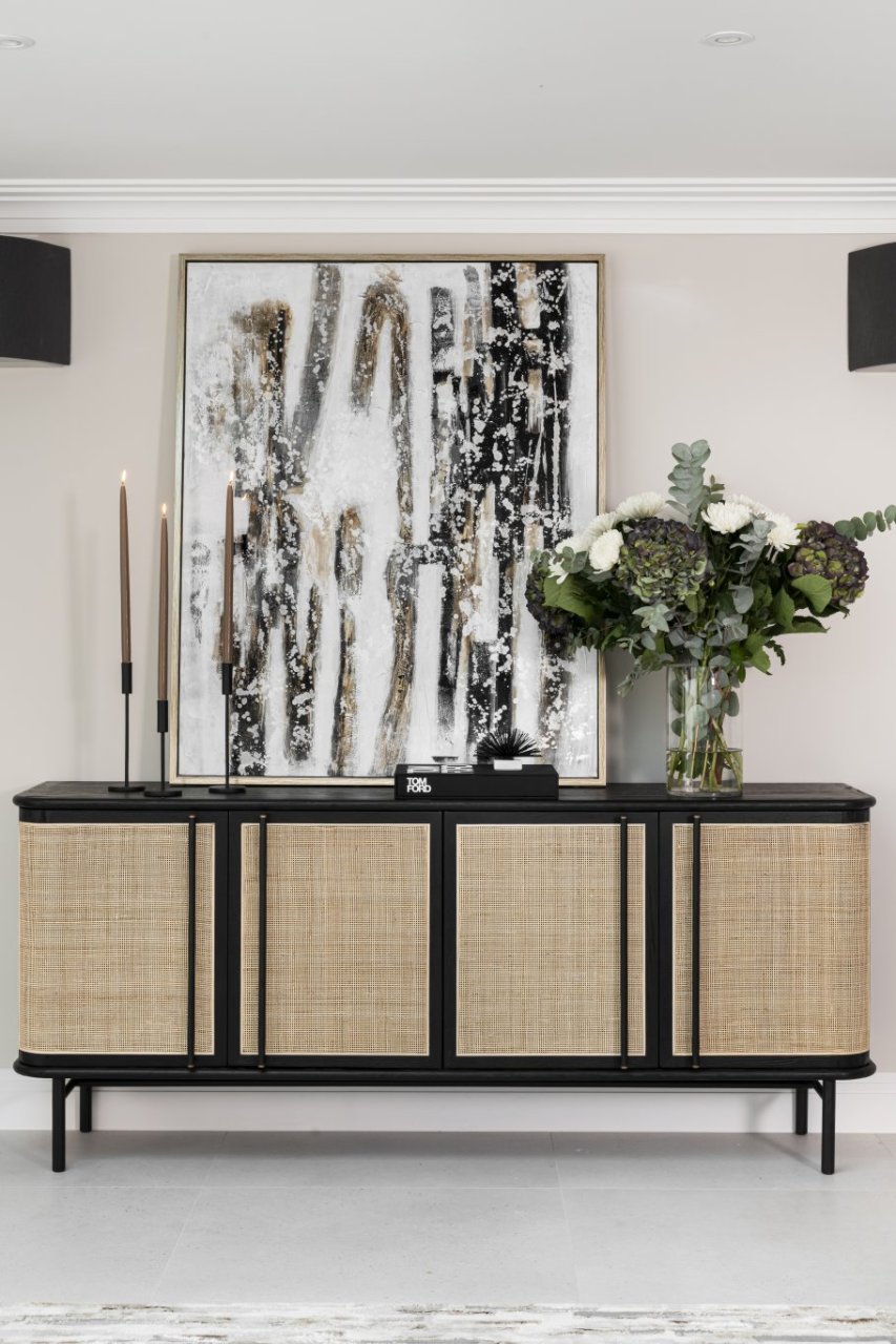 The TV unit is the main focus, showcasing its wide doors and intricate rattan facade. On the top it has a set of candles, wall art and an elaborate bouquet of flowers. 