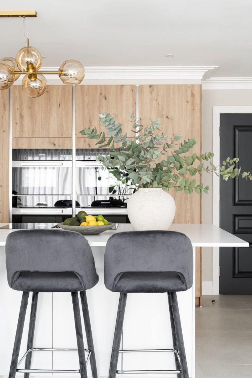 The side of an open plan kitchen island with two barstools facing it with their backs to the camera and a bowl of avocadoes, limes and lemons in front.