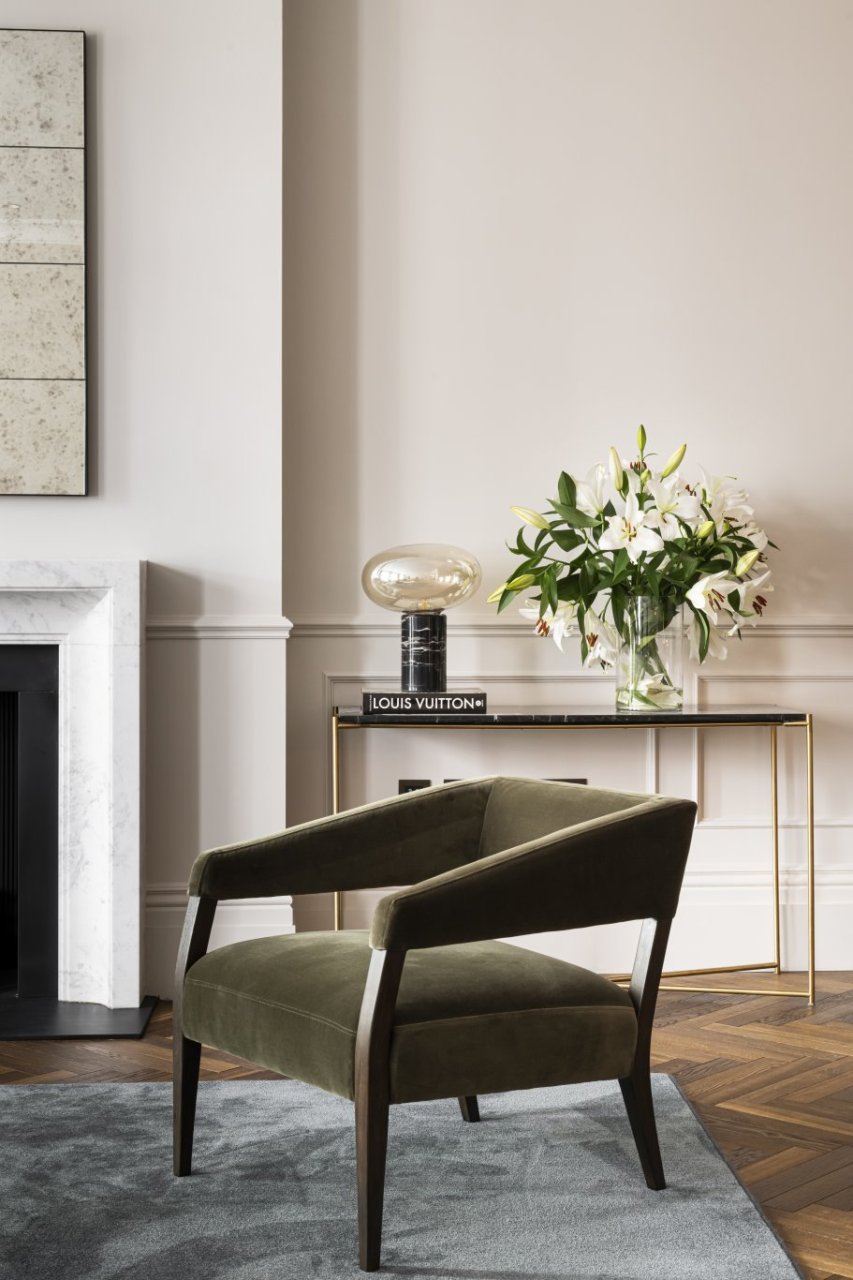Focus on a luxuriously upholstered armchair with rich, dark brown wooden accents placed in front of a console table with a lamp and bouquet of flowers on top. 