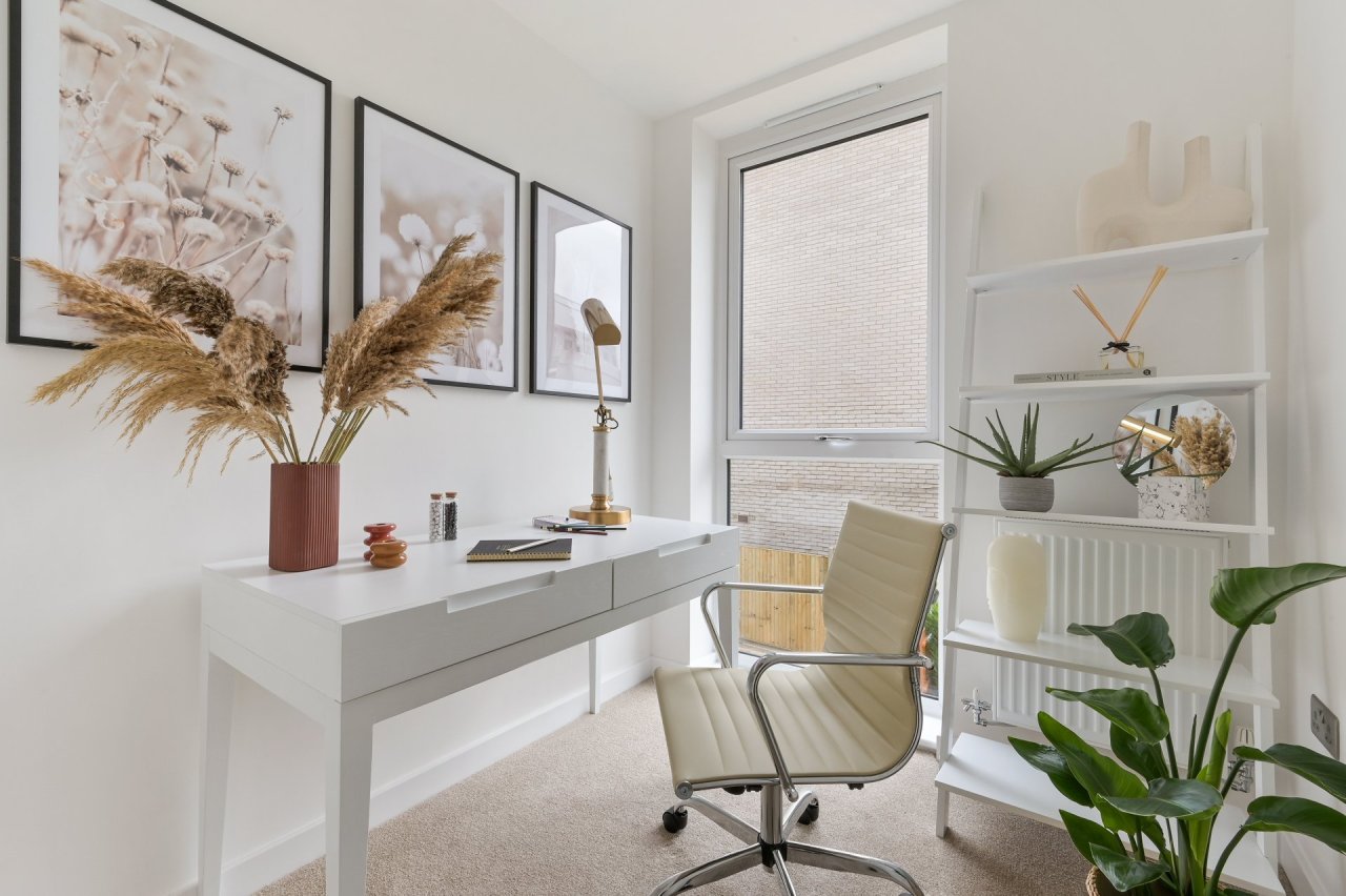 A bright home office with white walls and white wood furniture such as a desk with two drawers, a wall-leaning shelf and a cream curvy office chair on wheels.