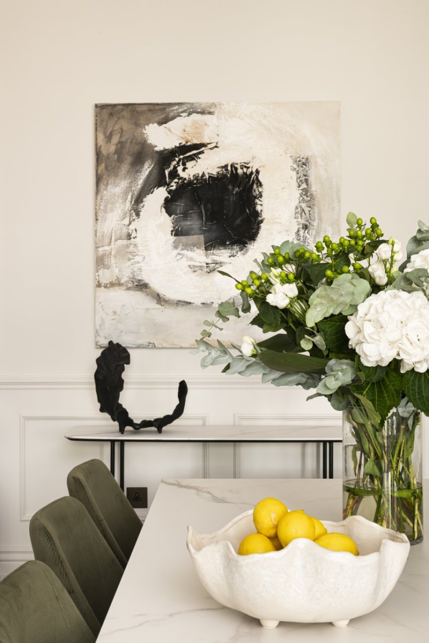 A view from the top of the table where you will find a vase of white flowers and a bowl of lemons. in the background a similar console table holds a horn-shaped abstract art and an abstract print above it.