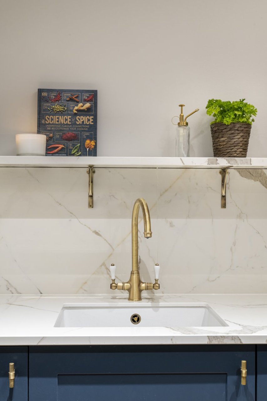 A kitchen sink with a brass, retro water tap. A spice book and fresh herbs are placed on a shelf above.