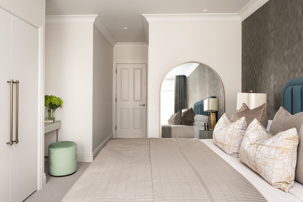 A view of the bedroom from inside the window. A large mirror sits beside the bed and highlights the cushions and bedside. At the foot of the bed there is a built in wardrobe next to a dressing table with a round teal pouffe.