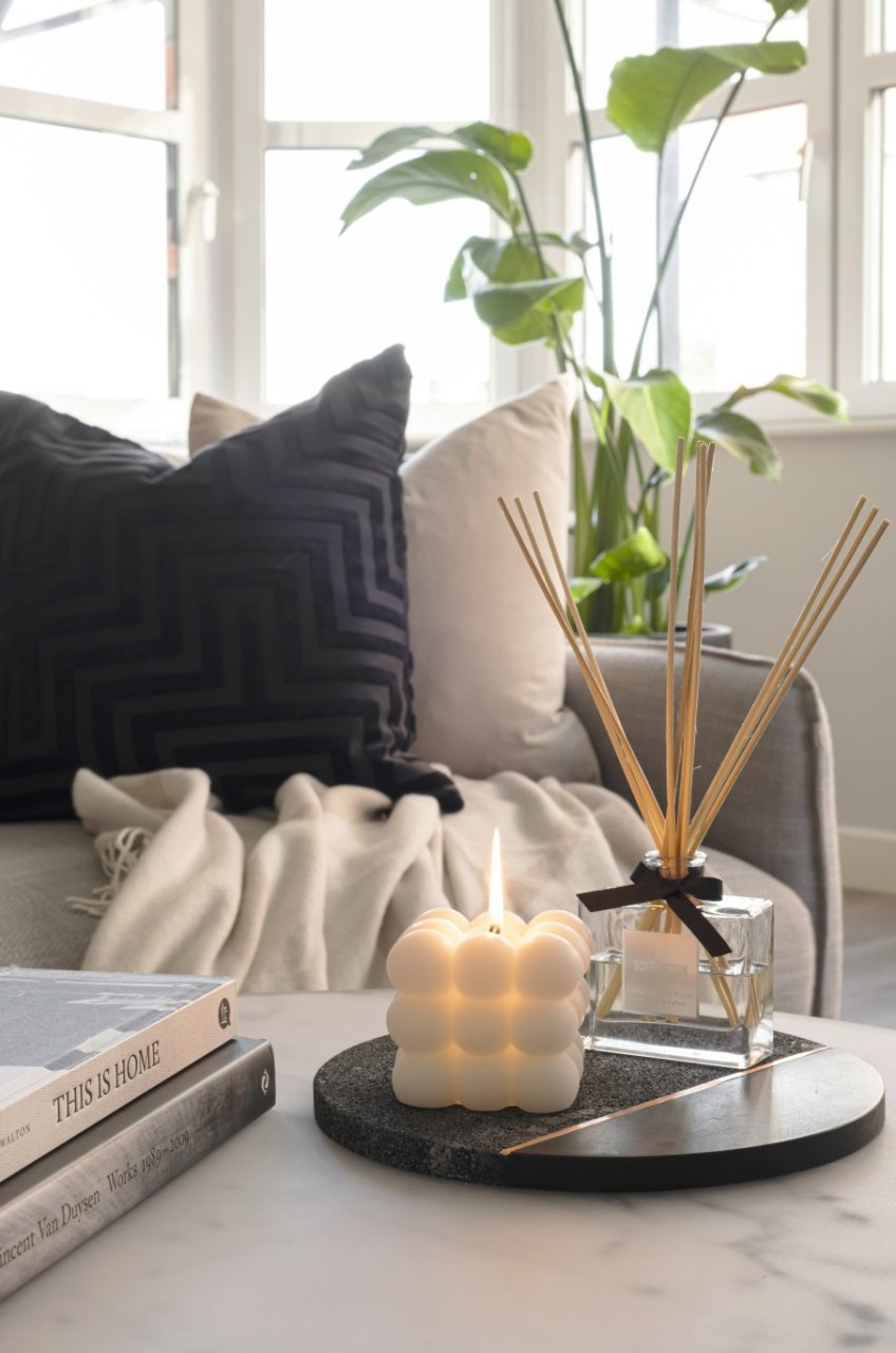 A focus on accessories professionally displayed on a coffee table. Two hardcover books sit next to a round platform that hosts a square candle and a cotton scent diffuser.