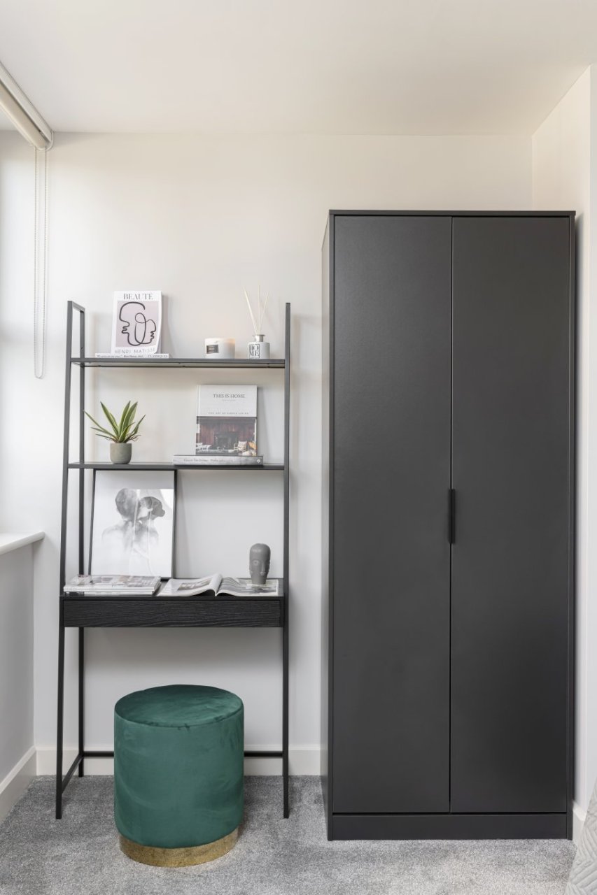A tall dark wardrobe stands on the right hand side of a nook. A dressing table with added shelves and accessories is placed beside it and a round stool underneath.