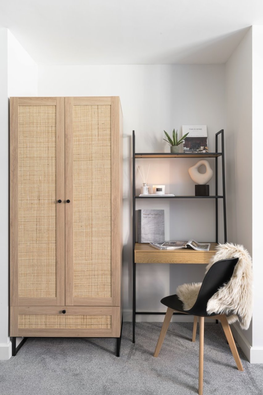 In a different nook of the same property there is a similar setup but with brighter, woody colours. A tall wardrobe with rattan accents and a bottom drawer sits next to a desk with storage accessories and a chair.
