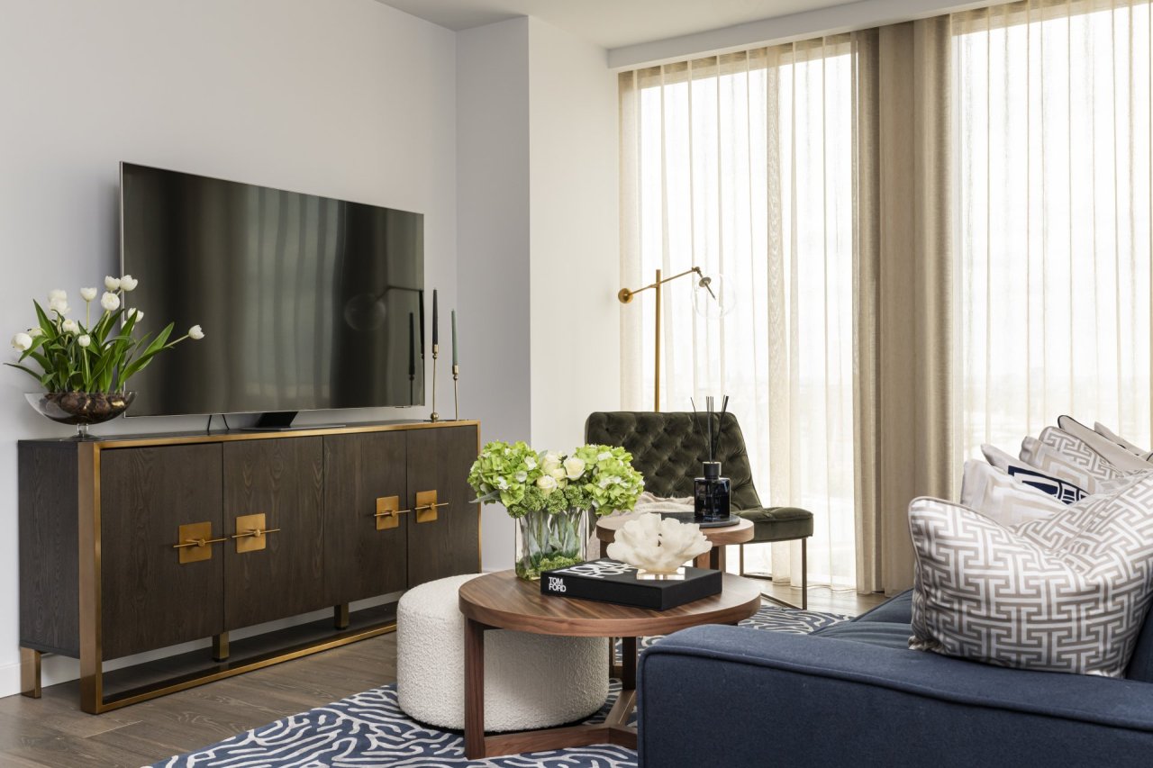 A 55 inch TV sitting on a TV unit inspired by safe vault design in deep wood and brass colours. Two round coffee/side tables and a boucle pouffe placed in front.