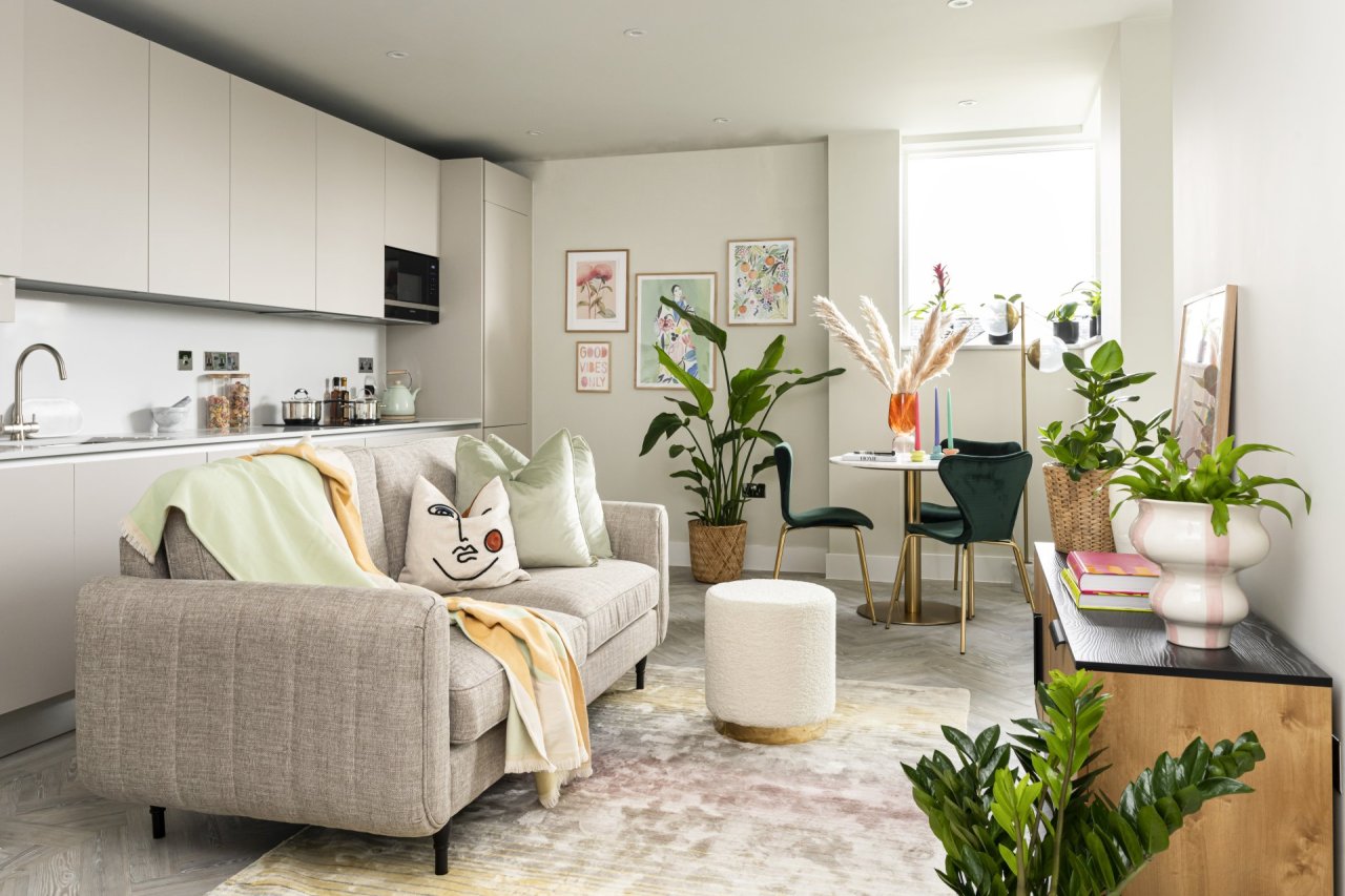 An open plan living room and kitchen, richly ornamented with green plants and colourful accessories. A 3 seater beige sofa faces a dressed TV unit. in the background a round table hosts 3 curvy upholstered dining chairs.