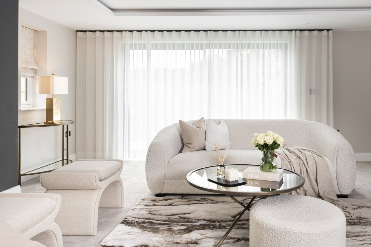 A room that is a heaven of boucle fabric and soft creamy colours. A bean-shaped-back sofa faces the camera over a round coffee table and a wide pouffe with two hourglass accent stools standing on the left side.