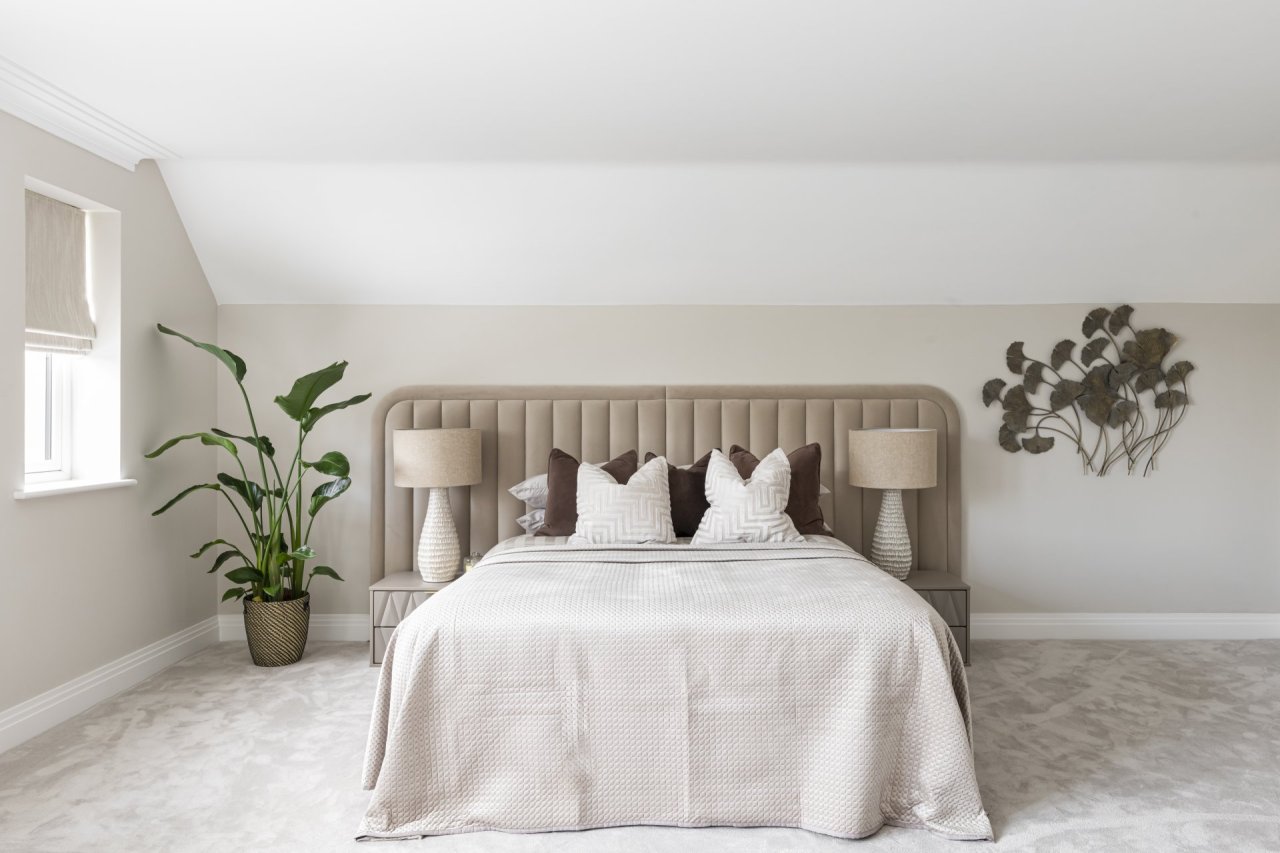 A view of a king size divan bed with an oversized cream-brown headboard and two symmetrical bedside drawers with slender lamps. The bedroom is spacious and features a plant on one side of the bed and a sculpture of leaves on the other.