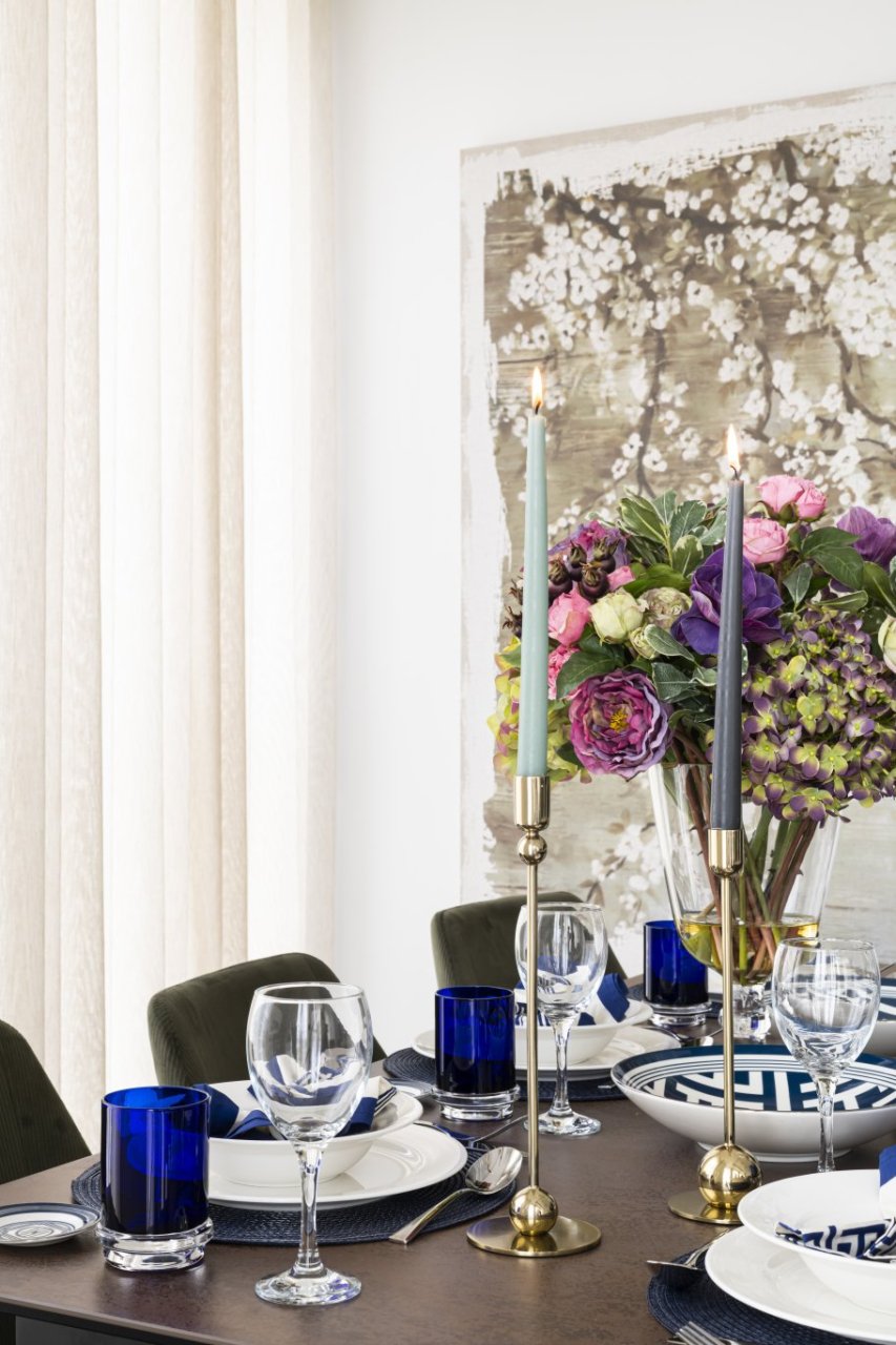 A beautifully set dining table with blue water glasses and expertly placed silverware, surrounded by green upholstered dining chairs. An intricate bouquet of flowers sits in front of a print of cherry blossom.
